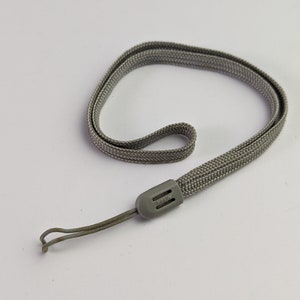 Pode incluir: Uma alça de pulso tecida em cinza para câmara com um design de cordão plano trançado e um deslizador de ajuste em plástico cinza. O deslizador apresenta a marca Nikon em relevo com letras claras, oferecendo um ponto de fixação seguro.