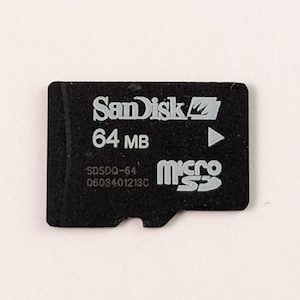 Może przedstawiać: Czarna karta pamięci microSD SanDisk z białym napisem SanDisk, 64 MB oraz logo microSD. Karta posiada również numer modelu SDSDQ-64 oraz numer seryjny 0603401213C nadrukowany w kolorze białym na przedniej powierzchni karty.