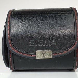 Puede incluir: Funda de cámara de cuero negro con costuras rojas. La funda tiene el nombre de la marca "SIGMA" grabado en relieve en la parte delantera. Un cierre de metal plateado asegura la funda. Diseñada para proteger una cámara o lente.