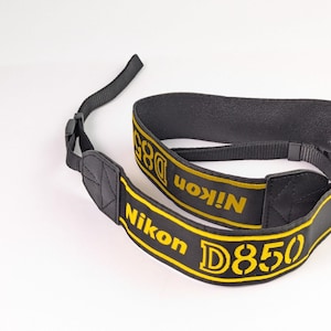 May include: A black camera strap featuring yellow embroidered text that reads Nikon D850. The strap includes adjustable black nylon webbing, a padded section with yellow border stitching, and plastic buckles for secure attachment to a camera body.