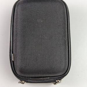 Puede incluir: Un estuche negro rectangular con cremallera y superficie texturizada. El estuche tiene la parte superior e inferior redondeadas, con un tirador de cremallera y bisagra plateados. Es probable que el estuche esté diseñado para proteger pequeños aparatos electrónicos u otros artículos.