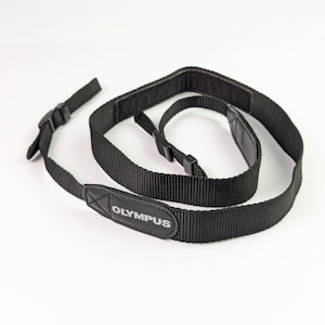 May include: A black camera neck strap featuring a woven nylon texture and a padded section. The strap includes adjustable plastic buckles and a black leather-like tab prominently displaying the white text OLYMPUS on the front surface.