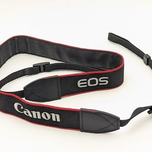 May include: Black Canon EOS camera strap with red trim. The strap features the Canon and EOS logos in white. The strap is made of a durable material and includes adjustable buckles for a secure fit.