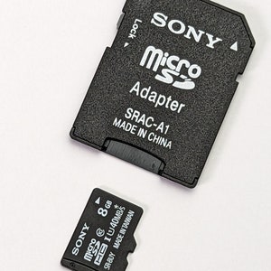 Op de afbeelding: Een zwarte Sony microSD-adapter met de tekst SONY, microSD, Adapter, SRAC-A1 en MADE IN CHINA, naast een zwarte 8GB Sony microSD-geheugenkaart met de tekst SONY, microSD, 8GB, 40MB/s, SR-8UY en MADE IN TAIWAN op het oppervlak gedrukt.