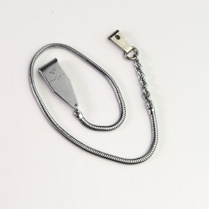 May include: A silver-toned metal chain with a snake-like texture, featuring a flat, tapered metal clip at one end engraved with the text GOSSEN and a small logo. The other end of the chain connects to a separate, smaller metal spring-loaded clip.