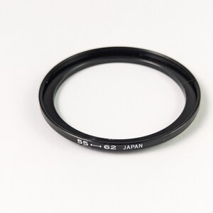 Puede incluir: Un anillo adaptador de objetivo de cámara circular de color negro con el texto blanco 55 <-> 62 JAPAN impreso en el lateral. El anillo de metal presenta un diseño roscado para acoplar filtros o accesorios de diferentes tamaños al objetivo.