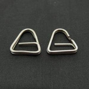 Può includere: Due fibbie triangolari in metallo color argento. Ogni fibbia ha una barra orizzontale al centro. Le fibbie sono esposte su uno sfondo scuro e strutturato.