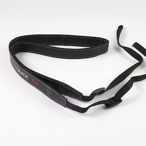 May include: A black camera neck strap featuring a padded section with white stitching and the text LUMIX G printed in white and red. The strap includes adjustable black nylon webbing ends with plastic buckles for secure attachment to a camera body.