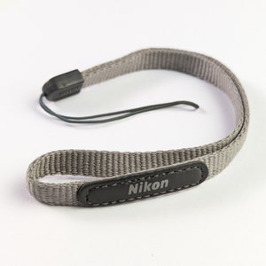 Op de afbeelding: Een grijze Nikon camerariem. De riem is gemaakt van geweven materiaal en heeft een zwart leren patch met het Nikon-logo. Een zwart plastic stuk en een zwart touwtje zijn aan de riem bevestigd.