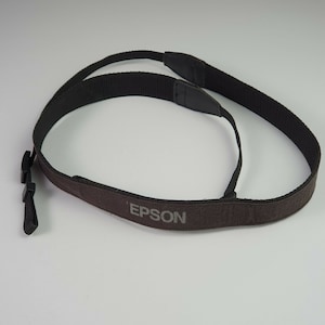 May include: A black and brown camera strap with the brand name "EPSON" printed in gray. The strap is made of a durable fabric and features adjustable buckles and a padded neck rest for comfort.
