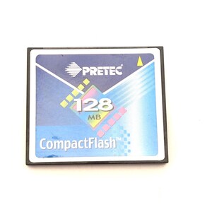 May include: A square Pretec CompactFlash memory card with a blue and white label. The label features the text PRETEC, 128 MB, and CompactFlash, along with a small yellow triangle and a graphic design of a square with colorful pixelated accents.