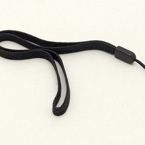 May include: A black wrist strap with a loop and a small plastic connector. The strap is made of a woven material and is designed to be worn around the wrist. The strap has a small loop at the end for attaching to a device.
