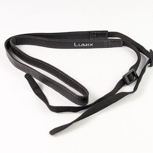 May include: A black camera neck strap featuring the white text "LUMIX" printed on a padded section. The strap is constructed from a combination of smooth material with grey stitching and adjustable black nylon webbing with plastic buckles.