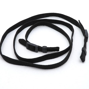 May include: A black woven nylon strap featuring a thin, flat design with adjustable plastic buckles and sliding keepers. The strap is coiled in a circular shape, showcasing the durable fabric texture and the functional black plastic hardware components.