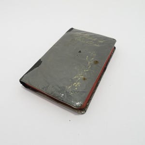 Peut inclure: Un livre rectangulaire avec une couverture gris foncé usée, ornée d'une inscription en lettres dorées et d'une délicate illustration florale dorée. Les tranches des pages sont teintées d'un rouge profond et la couverture est abîmée.