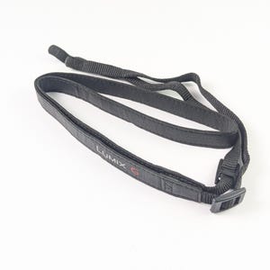 May include: A black camera neck strap featuring the text LUMIX G in white and red lettering. The strap is constructed from a textured material and includes adjustable black plastic buckles and thin nylon webbing connectors at the ends.