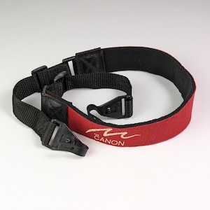 May include: A black and red camera neck strap with the word "Canon" embroidered in gold. The strap has adjustable black plastic buckles and a soft, padded red section for comfort.