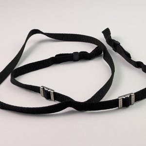 May include: A black camera strap made of woven nylon webbing featuring two silver-toned metal adjustment buckles. The strap includes small black plastic sliders for length modification and is designed with two attachment points at the ends.