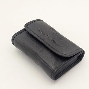 May include: A black rectangular camera case with a textured finish and a fold-over flap. The words Digital Camera are embossed on the front flap along with a small logo. The case features stitched edges and a compact, structured design.