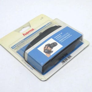Pode incluir: Uma alça de mão para câmera Hama preta embalada em um blister de plástico transparente sobre um suporte de papelão. A embalagem apresenta gráficos azuis e brancos, o logotipo Hama e o texto Patented air cell technology e Adapted tripod mount.