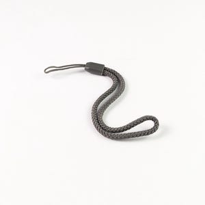 Pode incluir: Uma alça de pulso trançada cinza-escura com um laço de cordão fino em uma extremidade e uma peça de conexão de plástico sólido. A alça é feita de material trançado e projetada para ser fixada a pequenos dispositivos eletrônicos ou acessórios.