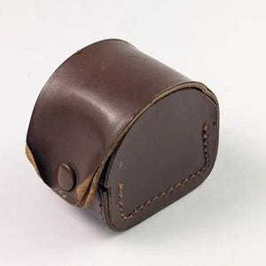 Puede incluir: Un estuche de cuero marrón con una parte superior redondeada y una base rectangular. El estuche tiene un cierre de botón y costuras visibles en los bordes. El cuero parece desgastado, lo que sugiere edad o uso.