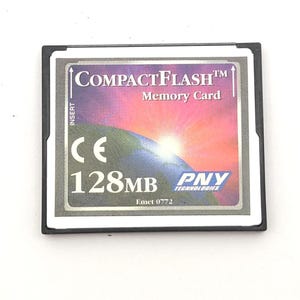 May include: A black PNY Technologies CompactFlash memory card with a 128MB capacity. The label features a sunrise over a globe graphic, the text CompactFlash Memory Card, CE, 128MB, PNY Technologies, and Emet 0772, with an arrow indicating insert.