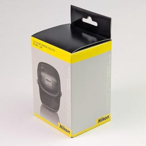 Può includere: Una scatola per custodia per fotocamera Nikon Coolpix. La scatola è principalmente grigia con accenti gialli e neri. La parte anteriore della scatola presenta un'immagine della custodia della fotocamera e il logo Nikon. Il testo sulla scatola include "ESTUCHE NIKON COOLPIX" e "DIGITAL CAMERA NIKON."