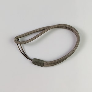 Pode incluir: Uma alça de pulso na cor taupe feita de material de cordão trançado. A alça possui um pequeno cursor retangular de plástico com acabamento fosco e a marca SONY em relevo, unindo as duas extremidades do cordão em um laço.