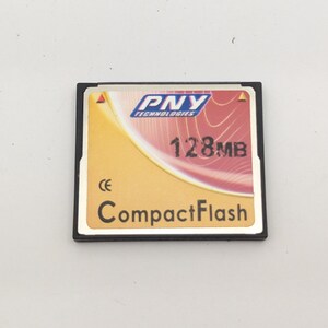 Puede incluir: Una tarjeta de memoria CompactFlash PNY Technologies de 128MB con un marco de plástico negro y una etiqueta dorada y roja con texto en blanco, el logotipo de PNY y una marca CE. La tarjeta tiene forma cuadrada con un borde metálico y pines.