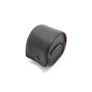 Könnte beinhalten: Ein schwarzes Lederetui mit gebogener Form, mit einem Knopfverschluss und einem Riemen mit einem Knopf. Das Innere scheint ein rotes Futter zu haben. Das Etui ist wahrscheinlich für ein kleines Objekt bestimmt. Maße nicht verfügbar.
