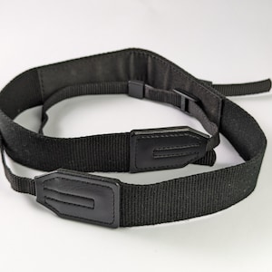 May include: A black camera strap made of woven nylon webbing featuring two reinforced faux leather end tabs with stitched detailing and adjustable plastic buckles for length modification, designed to securely hold a camera device.