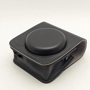 Può includere: Custodia per fotocamera in pelle nera con parte superiore arrotondata e base rettangolare. La custodia ha un interno a contrasto di colore chiaro e il marchio "CATUL" è visibile sulla parte superiore. La custodia è progettata per proteggere una fotocamera.