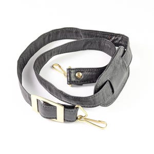 May include: Black leather bag strap with gold-colored metal hardware. The strap features a padded shoulder section and two swivel clasps for easy attachment. The hardware includes a rectangular buckle and two swivel clasps.