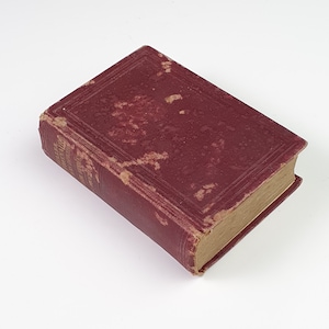 Op de afbeelding: Een klein, antiek boek met een versleten, bordeauxrode kaft. De kaft heeft een getextureerde uitstraling met zichtbare slijtage. De randen van het boek zijn verouderd en de pagina's zijn vergeeld. De titel is zichtbaar op de rug.