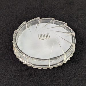 May include: A clear, circular lens filter with a textured, ridged edge. The word "HOYA" is printed in the center. The filter is set against a dark background.