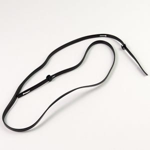 May include: A thin black adjustable strap featuring a smooth finish and a small circular fastener. The strap is flexible and curved, with visible slots along its length for adjustment, creating a simple and functional design for various uses.