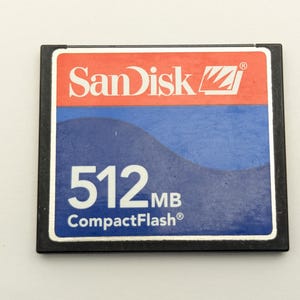 Può includere: Una scheda di memoria CompactFlash nera con un'etichetta rossa e blu. L'etichetta presenta il logo "SanDisk" e il testo "512MB CompactFlash". La scheda è rettangolare con angoli arrotondati.