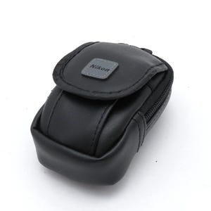 Pode incluir: Um estojo retangular preto para câmara com cantos arredondados e fecho de aba. A parte frontal da aba apresenta um emblema quadrado cinzento com a palavra Nikon impressa a preto. O estojo tem costuras pretas visíveis e um fecho de correr.