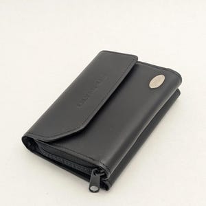 Puede incluir: Cartera de cuero negro con cierre de solapa y un emblema plateado. La cartera tiene un cierre de cremallera y la palabra "OLYMPUS" está grabada en la solapa. La cartera está cerrada.