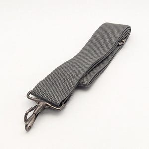 Peut inclure: Une sangle de remplacement en nylon tissé gris dotée d'un mousqueton pivotant en métal à une extrémité. La bande en tissu durable présente une surface texturée et est repliée sur elle-même, mettant en valeur la solidité de la construction.