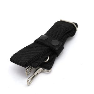 May include: A black nylon strap with metal swivel clasps and a metal D-ring. The strap has two black snap closures. The strap is designed to be used with a bag or other item.