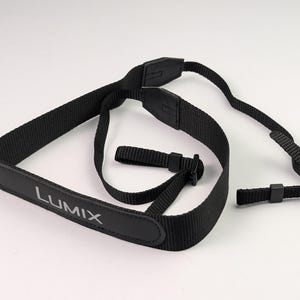 Puede incluir: Correa de cámara negra con la palabra "LUMIX" impresa en blanco. La correa está hecha de un material tejido y tiene hebillas de plástico ajustables para ajustar la longitud. La correa está diseñada para sujetarse a una cámara.