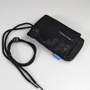 Pode incluir: Uma bolsa retangular preta com fecho de aba, apresentando um padrão floral tonal subtil e o texto { Urban Begz } impresso na frente. Inclui um cordão preto com um fecho de plástico ajustável e uma pequena etiqueta azul na lateral.