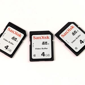 Pode incluir: Três cartões SD SanDisk pretos de 4 GB. Cada cartão tem o logotipo SanDisk em vermelho, as palavras "Video Buffer" e a capacidade de "4 GB" em um rótulo branco. Os cartões são retangulares com um canto entalhado.