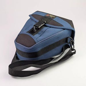 Könnte beinhalten: Eine blau-schwarze Kameratasche mit einem oberen Deckel, der durch eine schwarze Kunststoffschnalle gesichert ist. Die Tasche verfügt über ein rechteckiges Etikett mit dem Text Weekend Foto, einen verstellbaren schwarzen Schultergurt und ein strukturiertes Design.