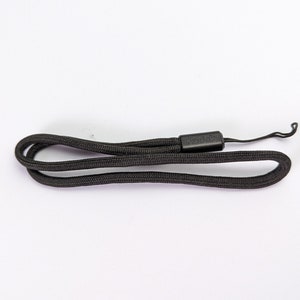 Pode incluir: Uma alça de pulso preta tecida para câmara com uma peça de conexão retangular de plástico com o nome da marca Kodak em relevo na superfície. A alça inclui um laço de cordão preto fino numa extremidade para fixação a um dispositivo.
