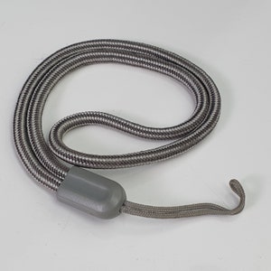 Pode incluir: Um cordão trançado cinza e branco com uma peça de conexão de plástico cinza liso. O cordão está disposto em forma circular e termina em um pequeno e fino laço cinza projetado para fixação em diversos objetos pequenos.