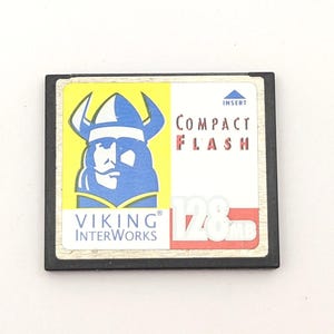 May include: A black rectangular CompactFlash memory card with a white label featuring a blue and yellow Viking logo. The label includes the text Viking InterWorks, Compact Flash, and 128MB in red and white. A small blue triangle indicates the insert direction.