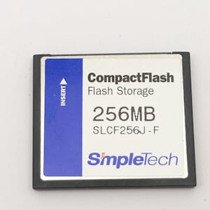 May include: A black and white SimpleTech CompactFlash memory card with a blue vertical stripe on the left side. The label displays the text CompactFlash Flash Storage, 256MB, SLCF256J-F, and SimpleTech, with an arrow pointing up labeled INSERT.